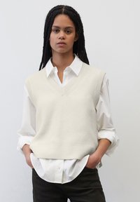 Hellbeige gestrickte Weste mit V-Ausschnitt, getragen über einem weißen Hemd mit Kragen, mit strukturiertem Stoff und lockerer Passform.