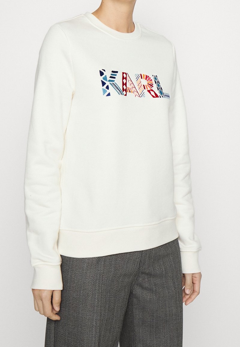 Personne portant un sweat-shirt blanc avec un texte coloré et à motifs « KARL » sur la poitrine et un pantalon gris texturé, debout devant un fond uni.