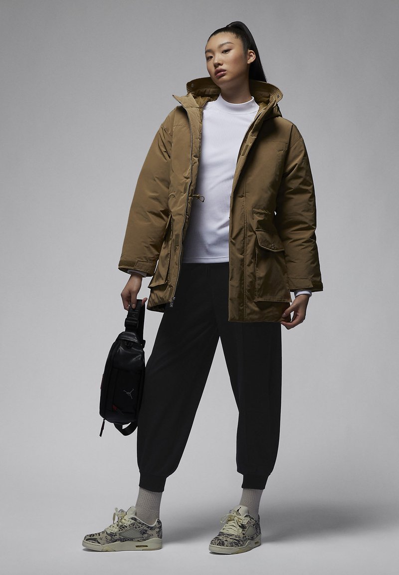 Chaqueta acolchada marrón con cremallera frontal, grandes bolsillos y bolsa ajustable. Top blanco de canalé, pantalones jogger negros y zapatillas con estampado, sosteniendo una bolsa.