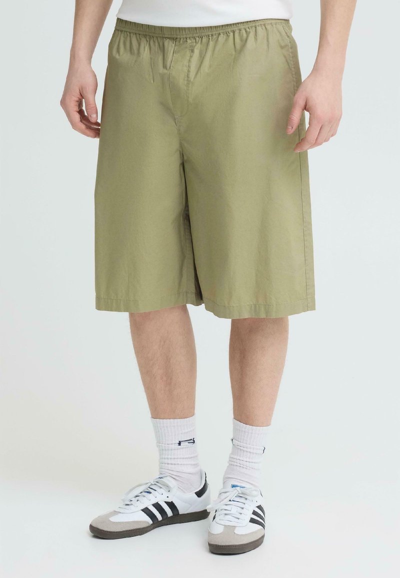 Homme portant un short kaki arrivant aux genoux, des chaussettes blanches montantes et des baskets blanches à rayures noires, se tenant sur un fond clair uni.