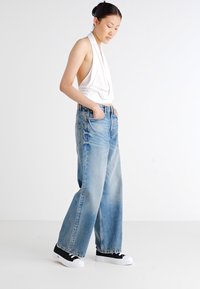 Jean en denim bleu clair à coupe large, associé à un haut sans manches blanc et des baskets noires avec des semelles blanches. Un look simple et décontracté.