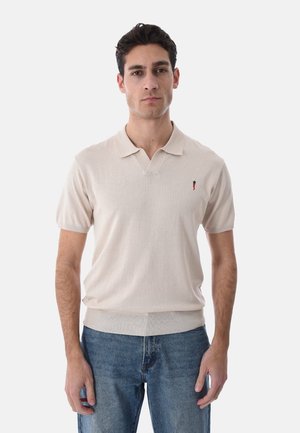 Uomo che indossa una polo beige a maniche corte con un piccolo logo a forma di peperoncino sul petto e jeans blu, in piedi davanti a uno sfondo bianco uniforme.