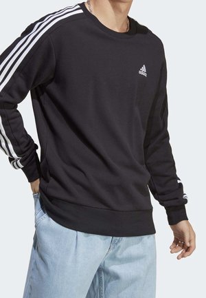 Homme portant un sweat-shirt noir Adidas avec des bandes blanches sur les manches et un jean bleu clair, debout avec une main dans la poche.