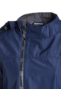 Marineblauwe windbreaker met een gestructureerde stof, verstelbare kraag en een rits met de merknaam "hummel." Inclusief maatlabels aan de binnenkant.