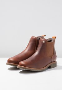 Botas de cuero marrón hasta el tobillo con punteras redondeadas, paneles elásticos en los laterales y un pequeño tacón. Forro suave y detalle decorativo en el hardware.