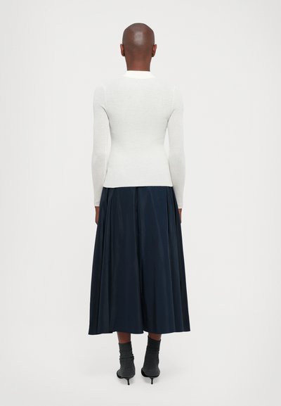Top bianco attillato a maniche lunghe con texture a coste, abbinato a una gonna lunga blu scuro fluida. Il look include stivali con tacco alti fino alla caviglia.