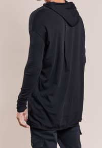 Musta pitkähihainen huppari rentolla istuvuudella, kevyestä kankaasta ja takana nappikiinnitys. Ominaisuuksiin kuuluu resorit hihansuissa ja pyöristetty helma.