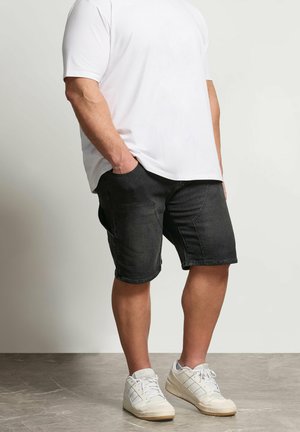 Mężczyzna ubrany w białą koszulkę, czarne szorty dżinsowe z kieszeniami i białe trampki, stoi z rękami w kieszeniach na szarej podłodze.