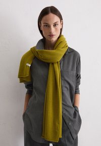 Donna che indossa una lunga sciarpa di maglia giallo senape avvolta sciolta intorno al collo e una camicia grigia a righe sottili con maniche arrotolate, mani nelle tasche.