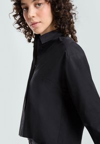 Czarna koszula z miękkiego materiału, z kołnierzykiem, zapięciem na guziki i skróconym fasonem. Gładka faktura z elegancką sylwetką.