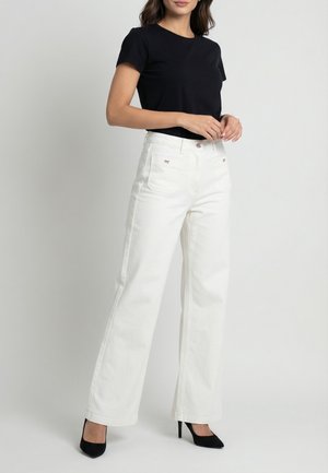 Femme portant un haut noir à manches courtes, un pantalon blanc taille haute à jambes larges, et des chaussures à talons noirs, debout les mains jointes.