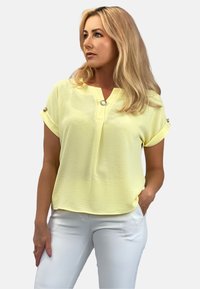 Żółta bluzka z krótkim rękawem, z dekoltem w kształcie litery V i dekoracyjnymi guzikami. Wykonana z lekkiego materiału, z podwiniętymi mankietami i luźnym krojem.