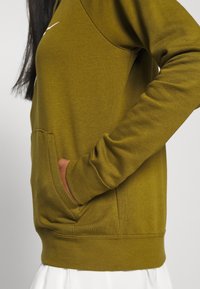 Person i en olivgrön sweatshirt med handen i framfickan, mot en enkel ljus bakgrund.