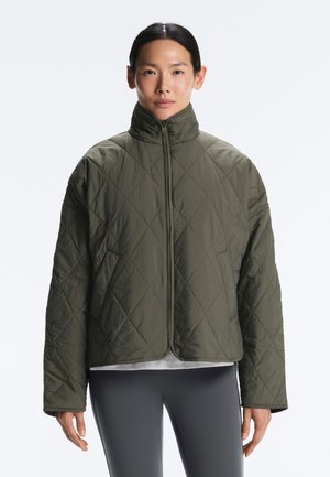 Kvinde iført en quiltet olivengrøn jakke med høj krave, stående med arme afslappet, mod en ensartet lys baggrund.