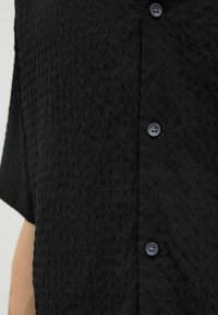 Camisa de manga corta negra texturizada con un cierre frontal de botones y un patrón sutil. Botón frontal en un tono oscuro a juego.