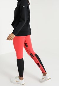 Černá mikina spojená s růžovými a černými legínami s bočním nápisem „NIKE“, s síťovanými prvky a bílými teniskami.