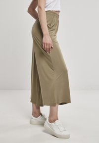 Kvinna iklädd olivgröna vida kapade byxor och vita sneakers som står på ett enfärgat vitt golv mot en ljus bakgrund.