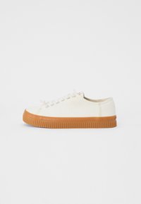 White canvas sneakers with a round toe and tan rubber sole. Laced design with a small red logo detail on the heel.