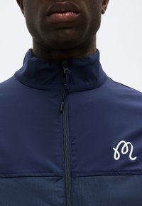 Κοντινό πλάνο στο κάτω μέρος του προσώπου και τον λαιμό ενός ατόμου που φοράει navy μπουφάν με φερμουάρ και λευκό κεντητό λογότυπο στο αριστερό μέρος του στήθους.