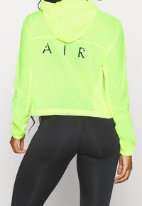 Veste courte légère de couleur jaune néon, avec des manches longues et une capuche. Présente le mot "AIR" imprimé en noir à l'arrière. Texture lisse, design sportif.