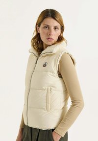 Chaleco de crema acolchado con un cuello alto y forro de borreguillo. Cuenta con una cremallera frontal y un bolsillo lateral. Lleva puesto sobre una camiseta de manga larga beige.