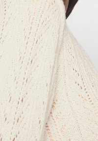 Weißer Strickpullover mit einem strukturierten Muster aus Zickzacks und offenen Maschen, der ein leichtes und luftiges Design zeigt.