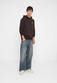 Hættetrøje i mørkebrun stof med en frontlomme, parret med vide jeans i blå og hvide sneakers, stående foran en ensfarvet baggrund.