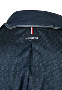 Schwarzes Jackeninnenfutter mit einem subtilen H-Muster in Dunkelblau. Enthält ein HECHTER-Label mit dem Wort "PARIS". Der Kragen hat eine kontrastierende Einfassung.