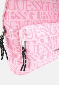 Rosa ryggsäck med stort mönster av "GUESS"-logotypen. Har en framficka med dragkedja, svarta dragskoaccenten och silverfärgad hårdvara.