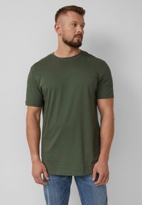 Olijfgroene katoenen t-shirt, ronde hals, korte mouwen, ontspannen pasvorm. Opvallend detail: klein logo op de borst. Gecombineerd met lichtblauwe jeans.