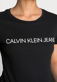 Černé bavlněné tričko s kulatým výstřihem, na přední straně má výrazný bílý text "CALVIN KLEIN JEANS". Měkká, hladká textura.