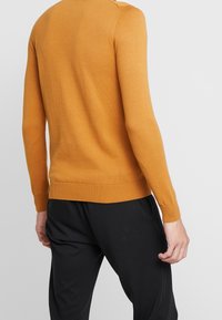 Suéter amarillo mostaza con cuello redondo, puños y dobladillo acanalados, y una textura suave. Combinado con pantalones negros, mostrando un look casual.