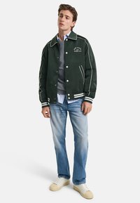 Grüne Bomberjacke mit weißen Paspeln, Frontdruckknöpfen und gesticktem Logo. Kombiniert mit einem grauen Pullover und hellblauen Jeans, abgerundet mit weißen Sneakers.