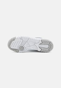 Egy fehér és szürke New Balance sneaker talpa, amelyen láthatók a talp mintázatai és a "Nem hagy nyomot" szöveg egy sima háttéren.