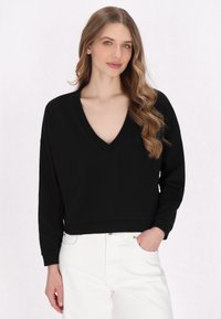 Pull-over noir à col en V avec des manches longues et un design court. Tissu doux et texturé ; associé à un pantalon blanc. Style simple et sans embellissement.