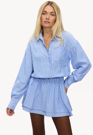 Femme blonde portant une robe à rayures verticales bleu et blanc à manches longues, avec une taille élastique et une poche poitrine, posant avec une main sur la hanche.