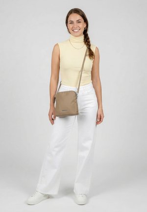 Young woman smiling, wearing sleeveless beige turtleneck, white wide-leg pants, white sneakers, and carrying a beige crossbody bag.