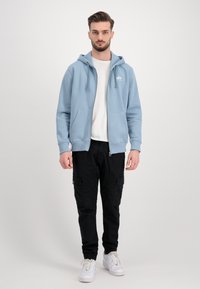 Felpa zip-up azzurra chiara con cappuccio regolabile, tasche frontali, indossata sopra una maglietta bianca, abbinata a pantaloni cargo neri e sneakers bianche.