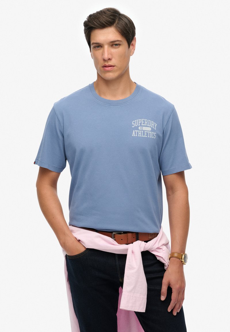 Camiseta de algodón azul con mangas cortas y cuello redondo. Cuenta con un texto gráfico blanco "SUPERDRY ATHLETICS" en el lado izquierdo del pecho.