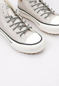 Sneaker alte in tela beige chiaro, con lacci neri, punta in gomma e striscia nera a contrasto sulla suola. Design testurizzato con dettaglio del logo.