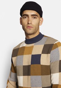 Strickpullover mit einem mehrfarbigen Karomuster in Braun, Creme und Marineblau. Hoher Kragen und gerippte Textur. Akzentuiert mit variierenden Streifenmustern.