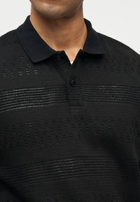 Homme portant un polo en maille noire avec un col à boutons et des motifs de rayures horizontales texturées sur la poitrine et les épaules.