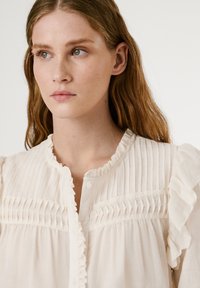 Weiße Bluse mit vertikalen Falten, Rüschensäumen und einer Knopfleiste. Aus weichem, leichtem Stoff mit strukturierter Oberfläche.