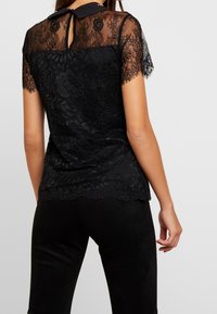 Mujer con una blusa corta de encaje negro con patrones florales y un escote en la espalda, combinada con pantalones ajustados negros.