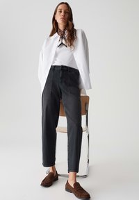 Femme portant une chemise blanche sur un haut blanc, un jean taille haute noir, des mocassins bruns et un foulard à motifs autour du cou, debout près d'une chaise.