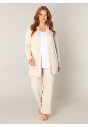 Vrouw met golvend kastanjebruin haar draagt een beige cardigan, wit topje, beige broek met wijde pijpen en beige sandaaltjes met bandjes, staand tegen een effen achtergrond.