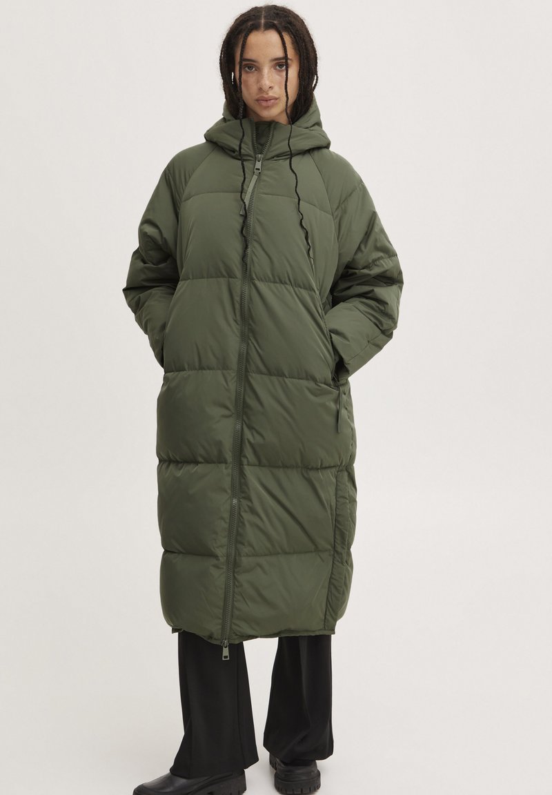 Grüne Pufferjacke mit Kapuze, ausgestattet mit einem Frontreißverschluss, im Oversize-Schnitt und mit gestepptem Design. Langes Modell mit Seitentaschen und gerippten Akzenten.