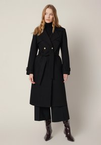 Czarny wełniany płaszcz trencher z paskiem w talii, podwójnym zapięciem i złotymi guzikami. Nosi się go na szerokie spodnie i błyszczące, wysokie botki.