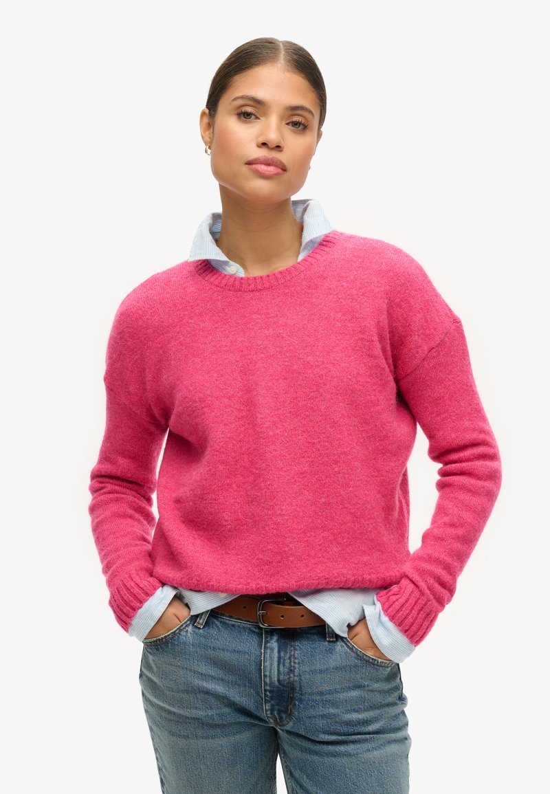 Różowy sweter dzianinowy z okrągłym dekoltem, zestawiony z jasnoniebieską koszulą z kołnierzykiem pod spodem. Stylizacja charakteryzuje się długimi rękawami i luźnym krojem.