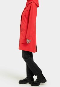 Röd vattentät jacka med huva, sidfickor och en avslappnad passform, kombinerad med svarta jeans med raka ben och klumpiga svarta stövlar.